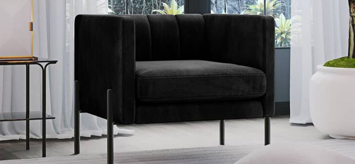 Modern black boxy armchair with channel-tufted sides and back, slender metal legs, positioned near a small side table and lamp, in a contemporary lounge setting.
