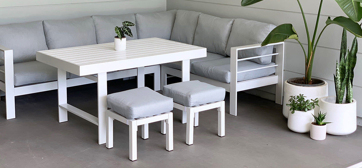 Minimal-style seating area with a white slatted-top table, L-shaped bench, and stools with gray cushions, surrounded by white planters with various houseplants.