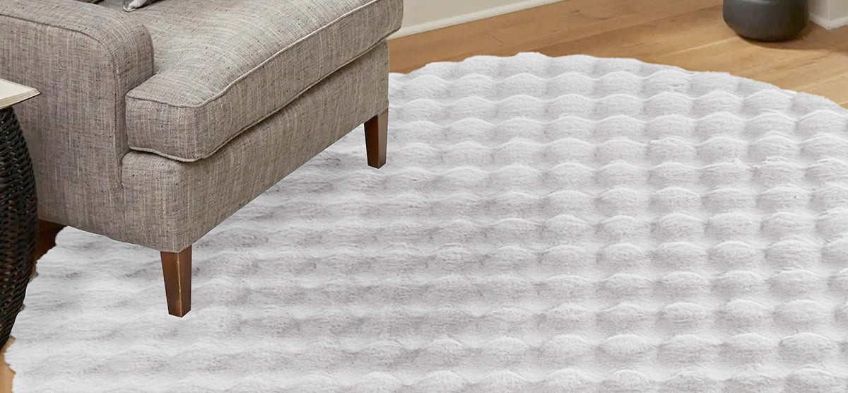 Mid-century modern gray upholstered sofa with wooden legs on light hardwood floor, accompanied by a round white plush area rug and a black woven side table.
