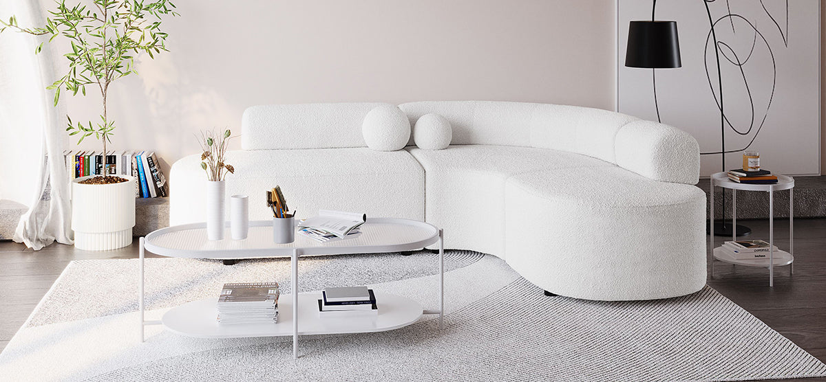A minimalist living room featuring a curved white sofa, oval coffee table with vases and books, potted plant, and abstract artwork, accented by dark flooring and soft daylight.