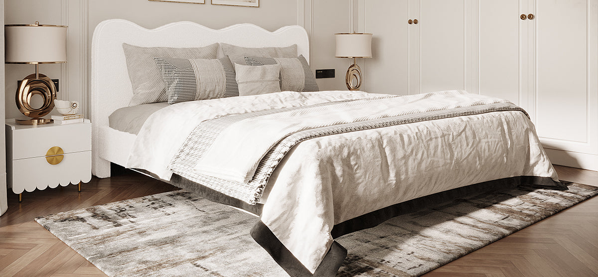 Neatly made bed featuring an off-white upholstered headboard, layered bedding, decorative throw, patterned rug, and gold-accent lamps on bedside tables in a contemporary bedroom.