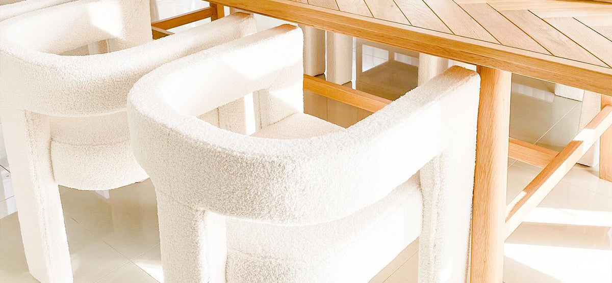 A light-filled dining area featuring a wooden parquet-style table and two modern white boucle-upholstered armchairs on a glossy cream-tiled floor with soft sunlight shadows.