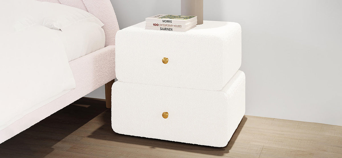Minimalist bedroom corner with a pale boucle-upholstered bed edge, white fabric-wrapped two-drawer bedside table with brass knobs, books, and lamp base. Light wood floor and plain white wall.