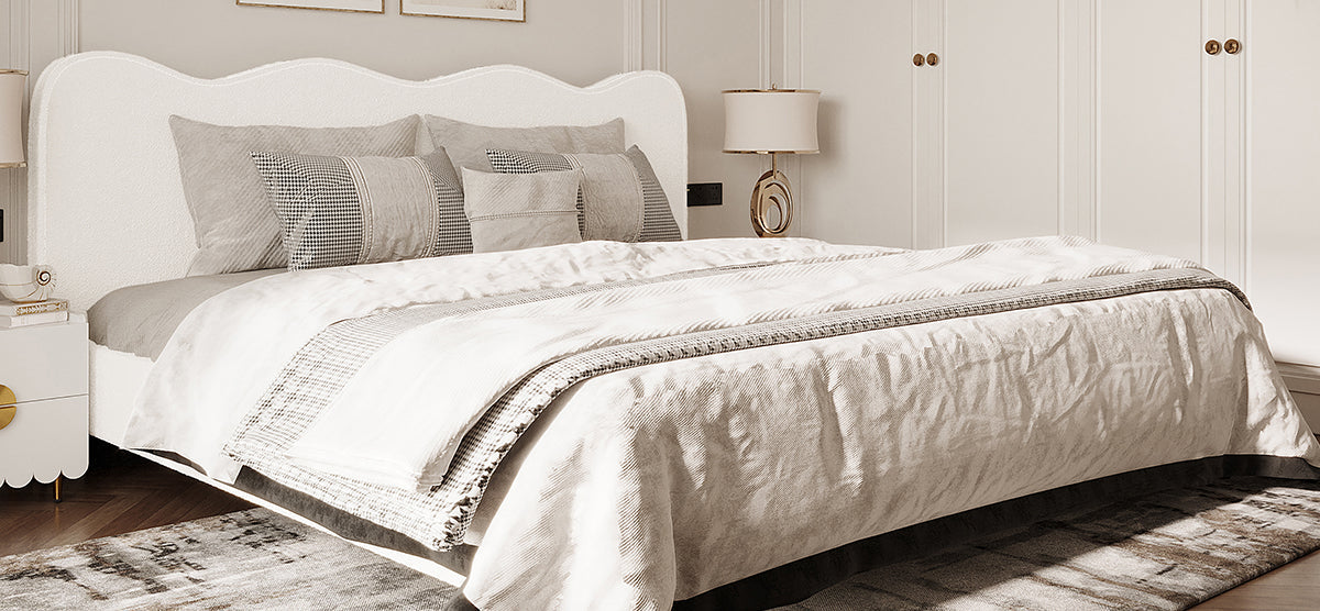 Neutral-toned bedroom with a scalloped headboard bed, layered gray and off-white linens, matching nightstands with lamps, and a patterned rug.