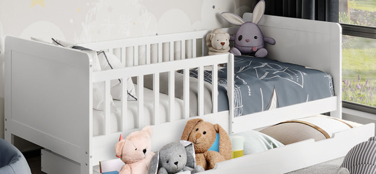 Children's bed with guard rails featuring blue nautical bedding, plush toys like a purple bunny and tan teddy bear. Pull-out trundle below holds additional toys and pillows.