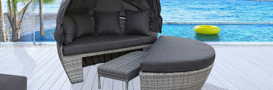 Semi-circular wicker daybed with dark gray cushions and pillows, accompanied by a curved ottoman and side table, set on a poolside deck with infinity pool view.