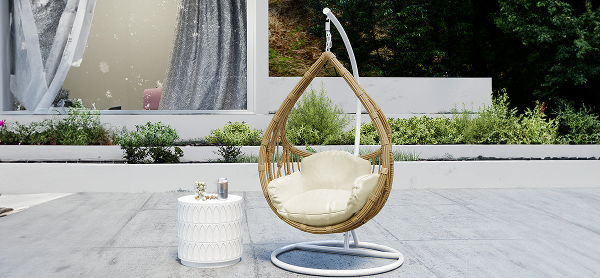 Teardrop-shaped rattan hanging chair with cream cushion on patio, adjacent to a white side table with a drink and snacks; surrounded by shrubs and greenery.