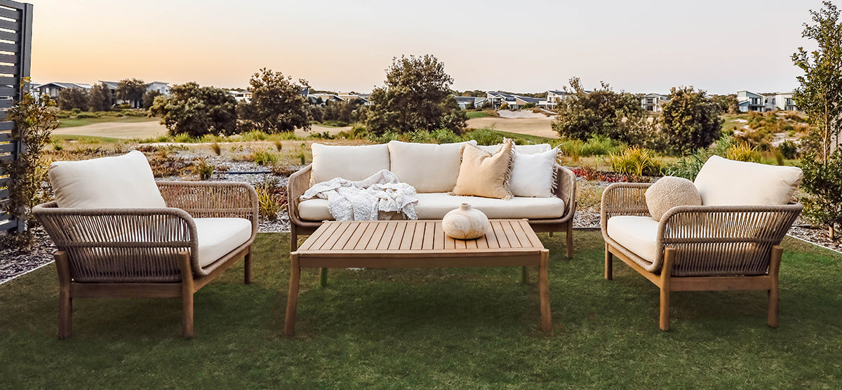 Outdoor lounge setting with a light-wood, rope-detailed sofa and armchairs, off-white cushions, and a slatted-wood coffee table on a lawn, under a soft sky.