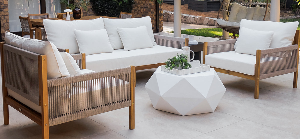 Modern outdoor seating area featuring a low, white-cushioned sofa and armchair with wooden frames and rope-woven sides, accompanied by a geometric coffee table with a pitcher and plant.