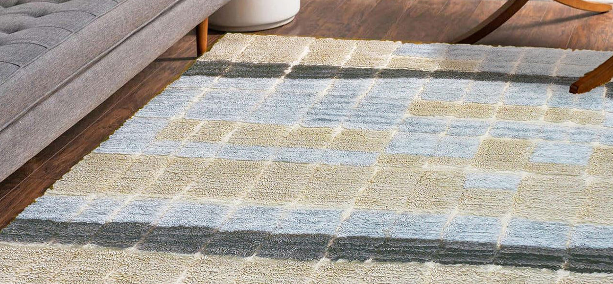 Checkerboard-style area rug in beige, gray, and pale blue on hardwood floor, with the edge of a gray sofa and a wooden chair leg visible.