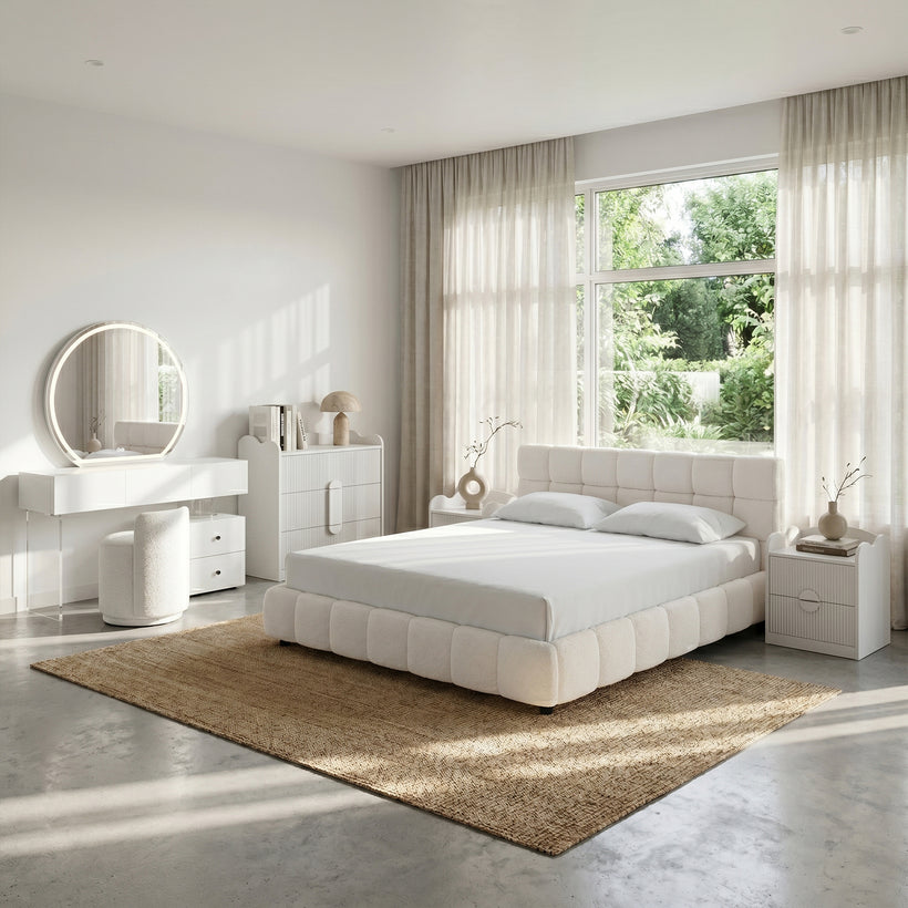Bright, contemporary bedroom featuring a white upholstered platform bed with a tufted headboard, matching white nightstands and dresser, illuminated round vanity mirror, textured beige area rug, floor-to-ceiling sheer curtains, and large windows overlooking lush greenery, creating a clean and serene atmosphere.