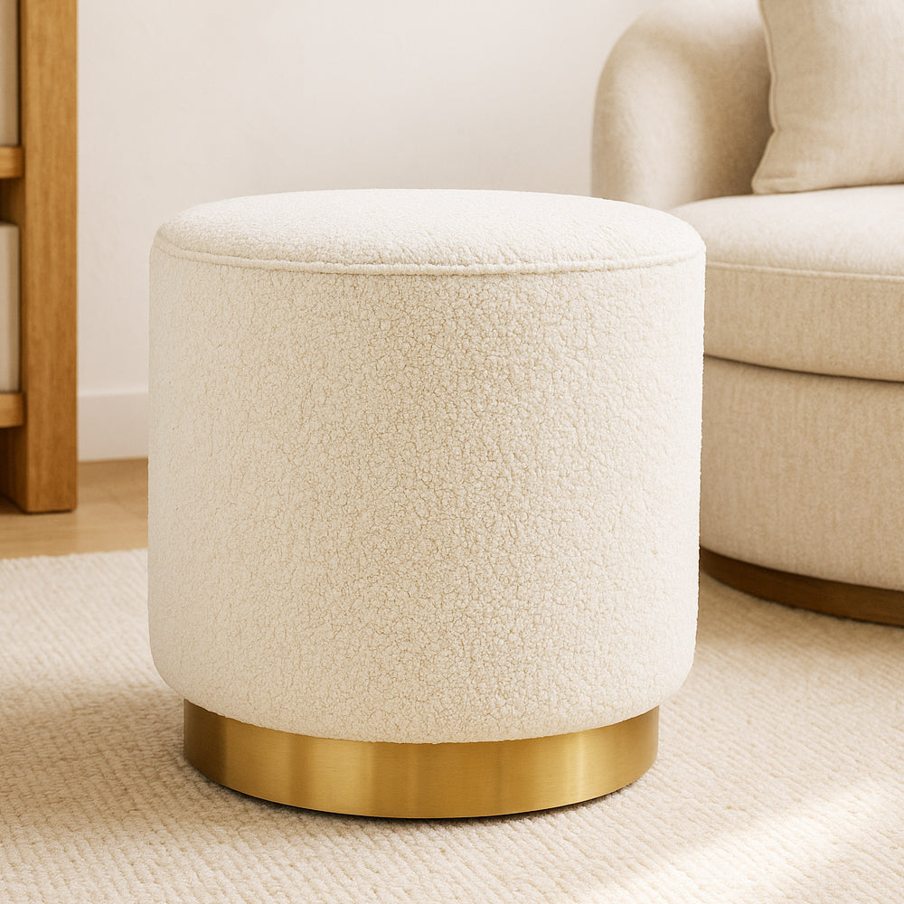 Angel Bouclé Ottoman in Cream White sits on a brushed brass-tone base, upholstered in cream bouclé fabric. It rests on a light rug, with a beige chair and wooden cabinet leg visible nearby.