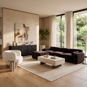 Dark brown velvet sofa with ottoman paired with a white bouclé armchair, travertine coffee table, and a black sideboard in a light-filled living room.