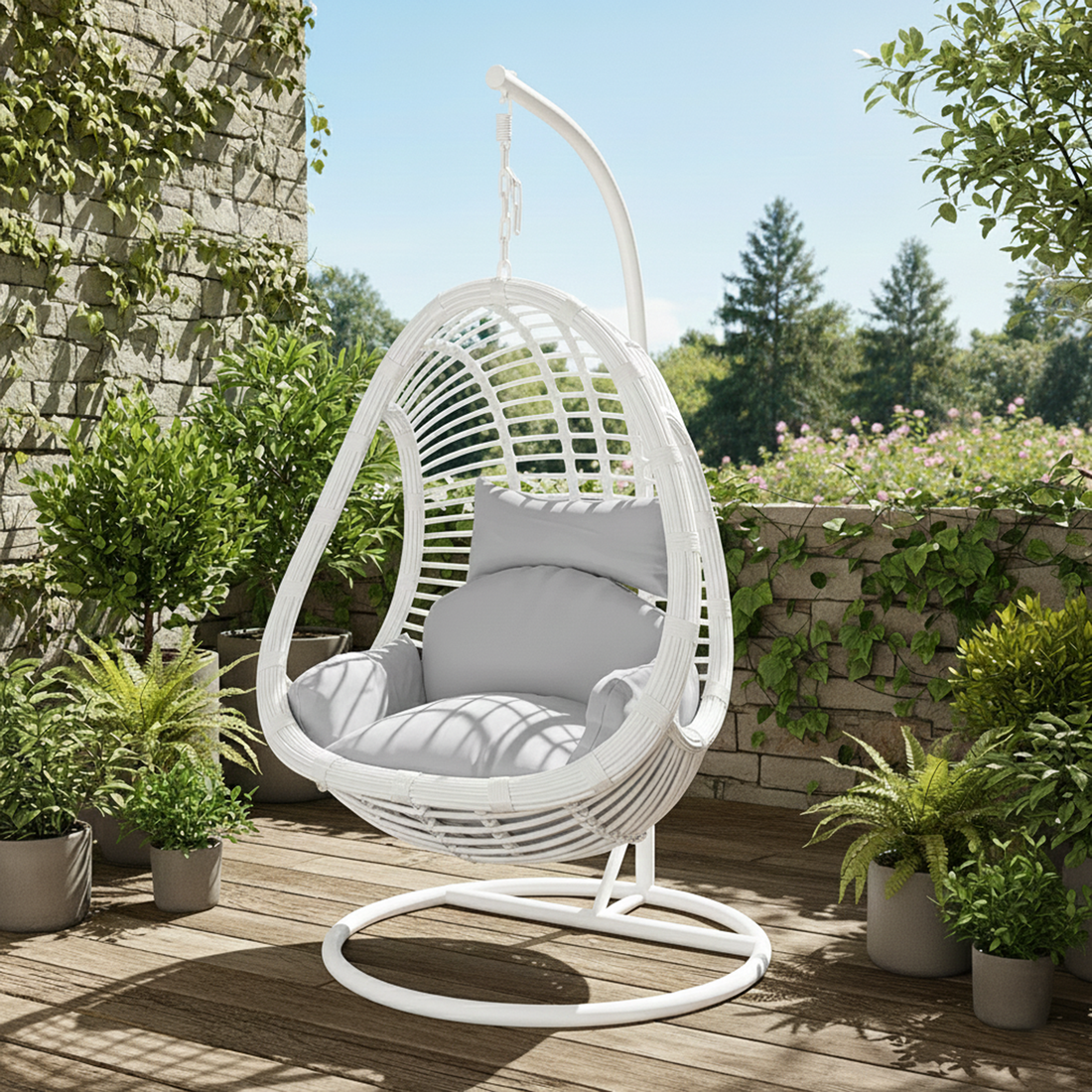 Deanna Curved Rattan Hanging Outdoor Egg Chair in White, featuring a woven egg-shaped design with plush cushions, hanging from a sturdy metal stand amidst a lush garden setting with potted plants.