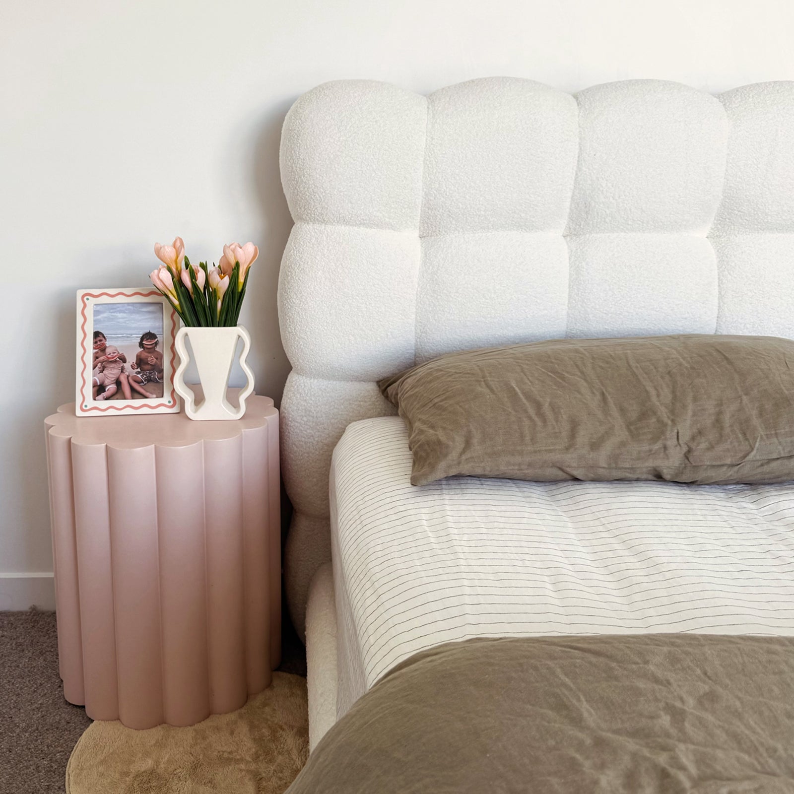 Aina Queen Boucle Bed Frame in Cream White with a plush tufted headboard, complemented by olive-brown linens and a nearby scalloped-edge pink side table with decorative accents.