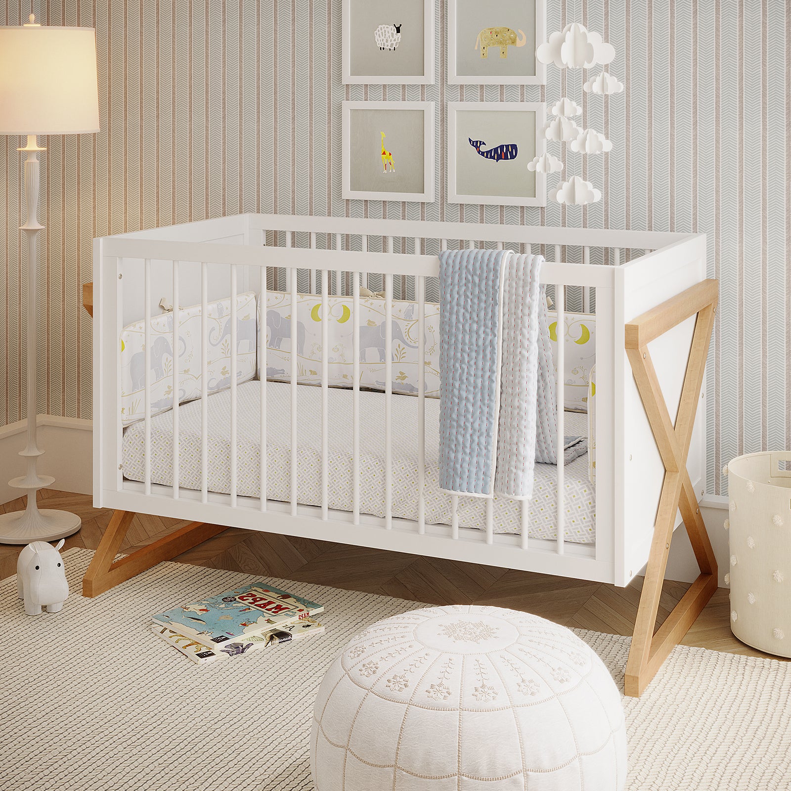 Aldwin Solid Wood Adjustable Baby Cot in White & Natural features slatted sides, X-style legs, and elephant-patterned bedding, with animal prints and a cloud mobile above, creating a cozy nursery setting.
