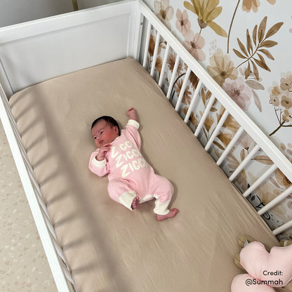 Aldwin Pine Adjustable Baby Cot - White/Natural, featuring a white-slatted design, modern X-leg base, and beige fitted sheet, situated against a wallpapered floral backdrop in a cozy nursery setting.