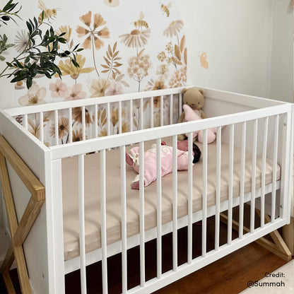 Aldwin Pine Adjustable Baby Cot with white slats and X-shaped supports, featuring a beige sheet, pink infant, teddy bear, and star cushion against floral wall decals, creating a cozy nursery setup.
