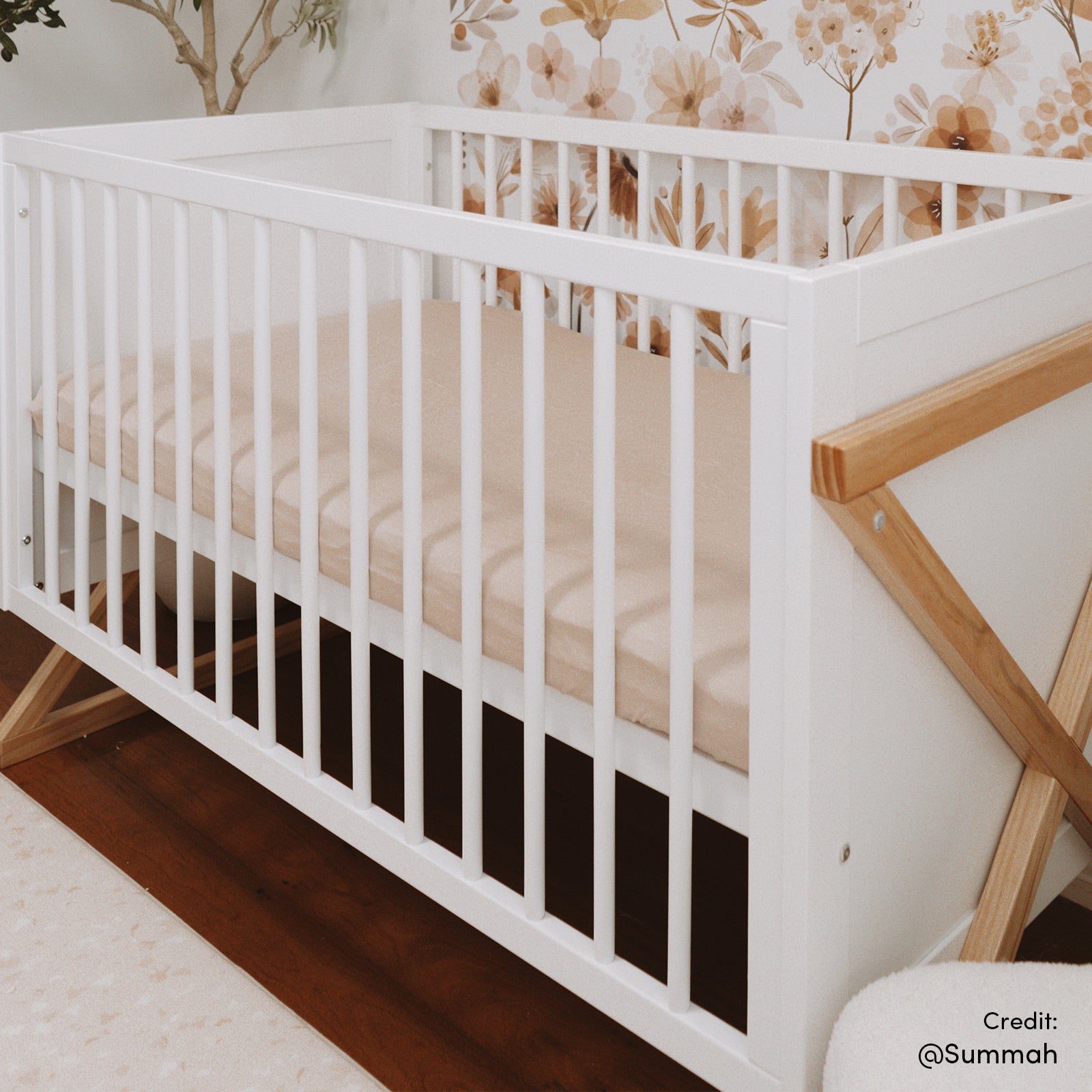Aldwin Pine Adjustable Baby Cot - White/Natural with slatted sides, X-style legs, and a beige fitted sheet, set against floral wallpaper in a nursery.