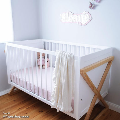 Aldwin Pine Adjustable Baby Cot - White/Natural features a white crib with X-shaped wood legs, pale pink sheet, plush toys, and a cream knit blanket in a bright, minimalist nursery setting.