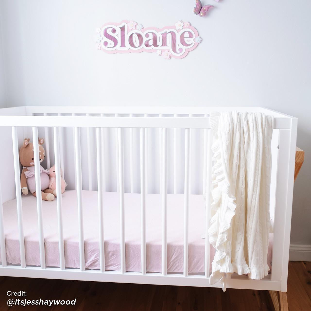 Aldwin Pine Adjustable Baby Cot - White/Natural in a nursery, features a white crib with a pale pink sheet, plush toys, a cable-knit blanket, and a decorative Sloane sign above.