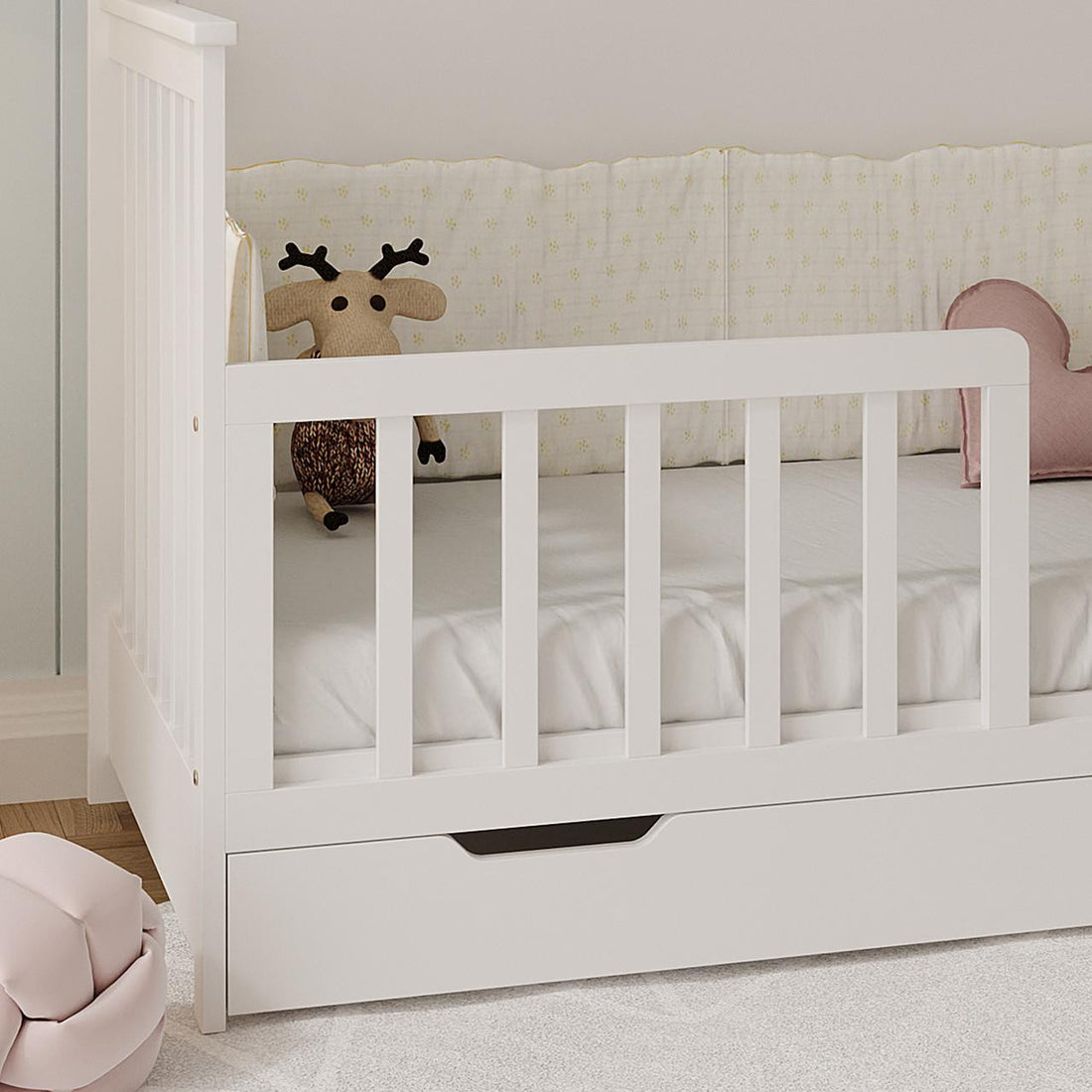 Azeem & Giro Baby Cot with built-in drawer, featuring Gydas 10cm Nursery Mattress. Includes a white sheet, patterned bumper, knitted reindeer toy, pink heart cushion, and nearby pink pouf.