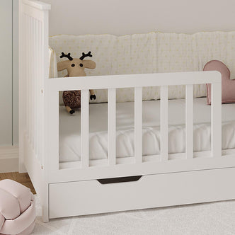 Azeem & Giro Solid Pine Baby Cot with Convertible Rail Set White in a nursery, featuring a plush reindeer toy, heart cushion, pull-out storage drawer, and blush-pink knot pillow on the floor.