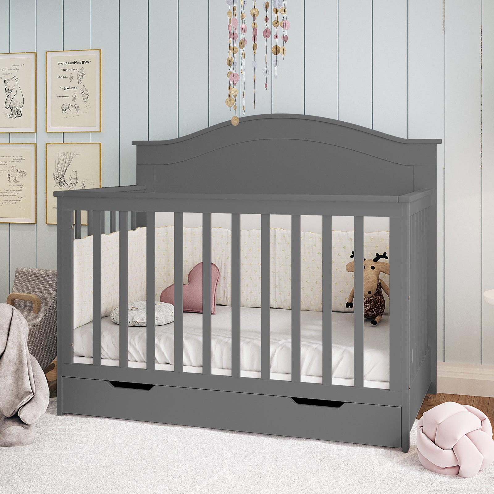 Azeem 3-in-1 Adjustable Solid Pine Baby Cot in Grey, featuring slatted sides, a pull-out storage drawer, star-patterned bumpers, heart and floral cushions, and a plush moose toy.