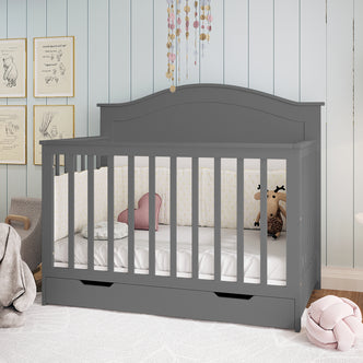 Azeem 3-in-1 Adjustable Solid Pine Baby Cot in Grey, featuring slatted sides, a pull-out storage drawer, star-patterned bumpers, heart and floral cushions, and a plush moose toy.
