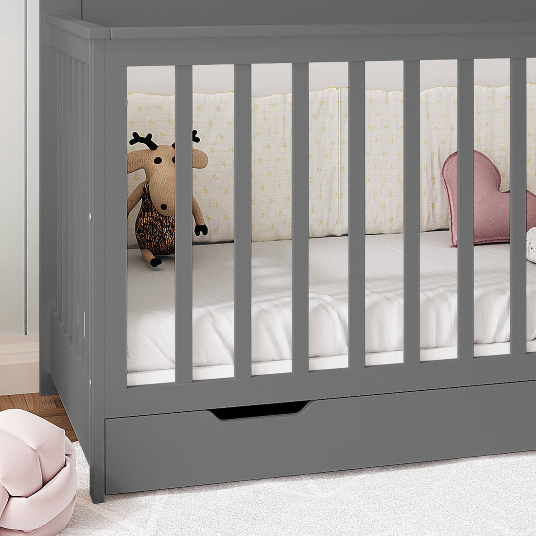 Azeem 3-in-1 Adjustable Solid Pine Baby Cot Grey with vertical slats, white fitted sheet, knitted moose toy, heart-shaped pink cushion, and built-in drawer, showcasing practical nursery style and functionality.
