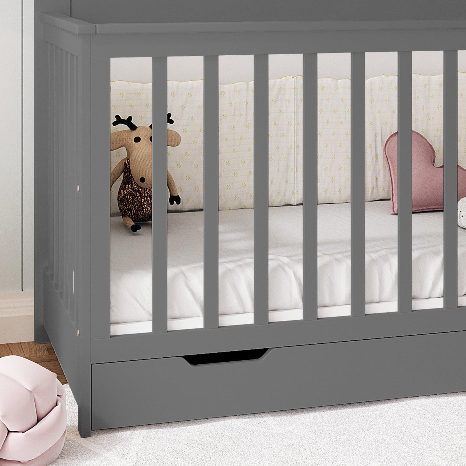 Azeem 3-in-1 Adjustable Solid Pine Baby Cot Grey with vertical slats, white fitted sheet, knitted moose toy, heart-shaped pink cushion, and built-in drawer, showcasing practical nursery style and functionality.