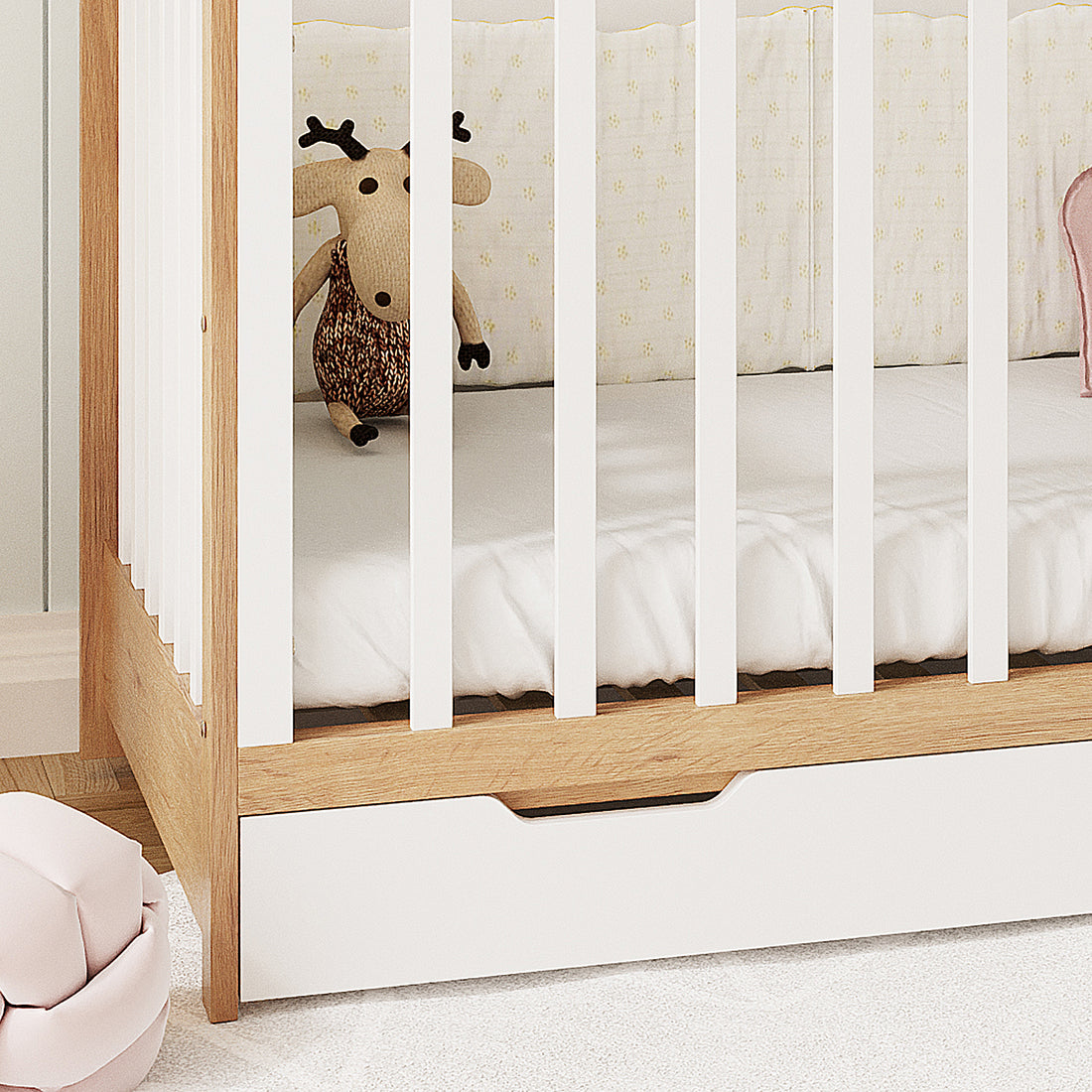 Azeem 3-in-1 Adjustable Solid Pine Baby Cot in White Natural, featuring light wood with white slats, a white mattress, pale bumper pad, built-in storage drawer, and a knitted deer stuffed animal.