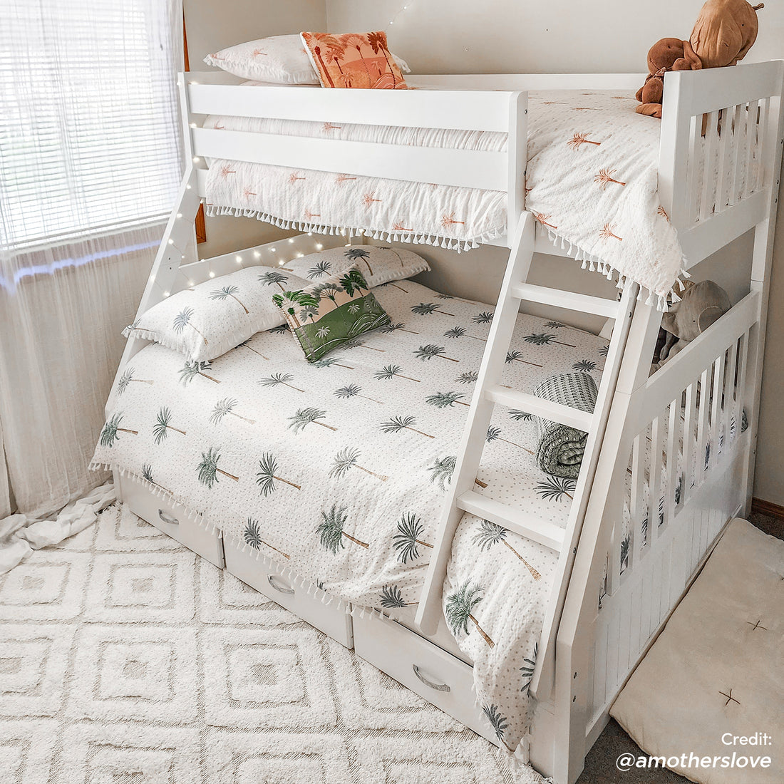 Cornelia Solid Wood Triple Bunk Bed with Storage in White, featuring a twin-over-double layout, palm-tree motif bedding, fairy lights, plush toys on top bunk, and built-in storage drawers below.
