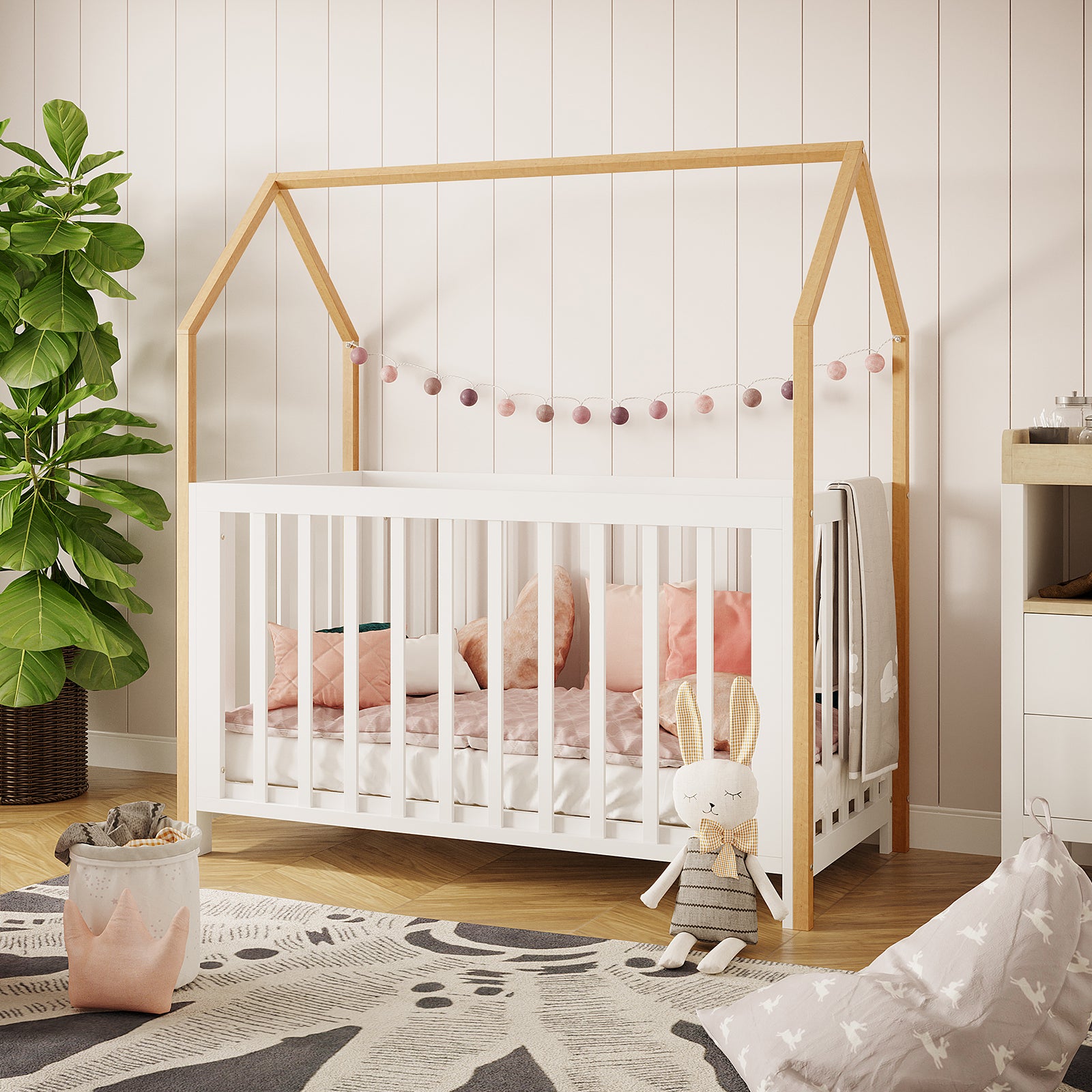 Damaris Adjustable Solid Wood Baby Cot in White & Natural, featuring a house-shaped frame with pastel felt garlands, soft bedding, and a cozy nursery setup with a plush bunny and decorative basket.