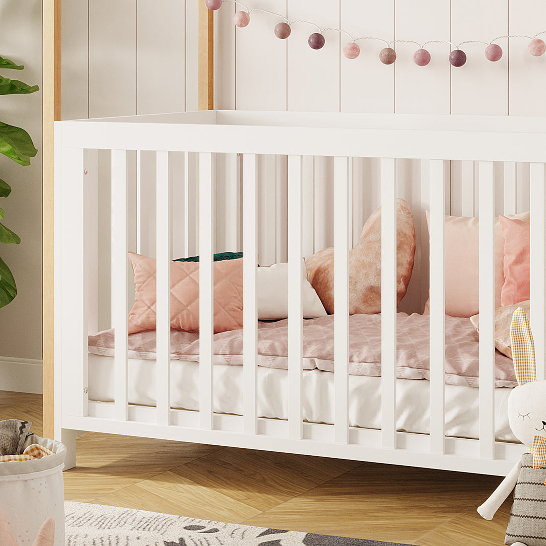 Damaris Solid Pine Baby Cot Bed - White/Natural features a house-shaped frame, white wooden structure with blush-toned cushions, and a pink garland above, creating a dreamy nursery atmosphere.