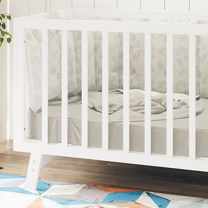Eudora Classic Solid Pine Adjustable Nursery Cot – White, featuring slatted sides, a grey mattress with a light blanket, and softly patterned crib bumpers, set on a wood-toned floor with a colorful rug visible.