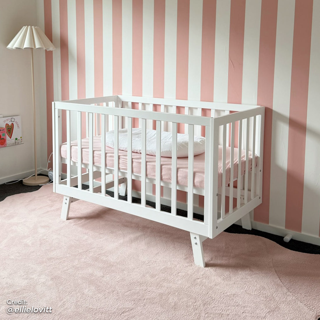 Eudora Classic Solid Wood Baby Cot in White with clean lines and pale pink mattress, set in a modern nursery with striped walls, a plush rug, and a paper-shade floor lamp.