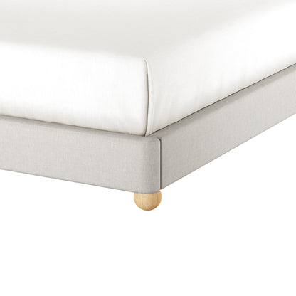 Close-up of the Ginnie Linen Curved Queen Bed Frame - Grey, showcasing the light-gray upholstered frame with a rounded wooden leg, and a mattress covered with a fitted white sheet.
