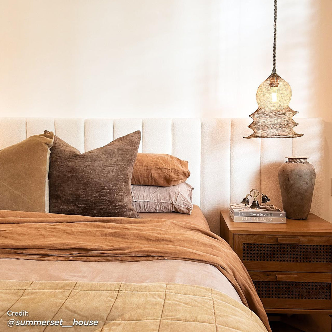 Aquila King Bouclé Extra Wide Headboard in Cream White centered in an earth-toned bedroom, accented by layered linens, velvet pillows, and a rustic nightstand with decorative items and a pendant light.
