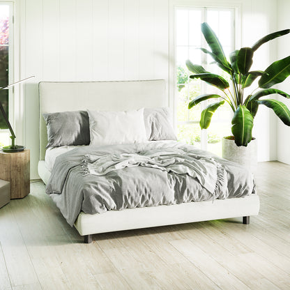Havvan Double Linen Upholstered Fabric Bed Frame - Off White features a soft headboard with a light fabric finish, paired with gray bedding in a serene bedroom setting with natural light and tropical plant accents.