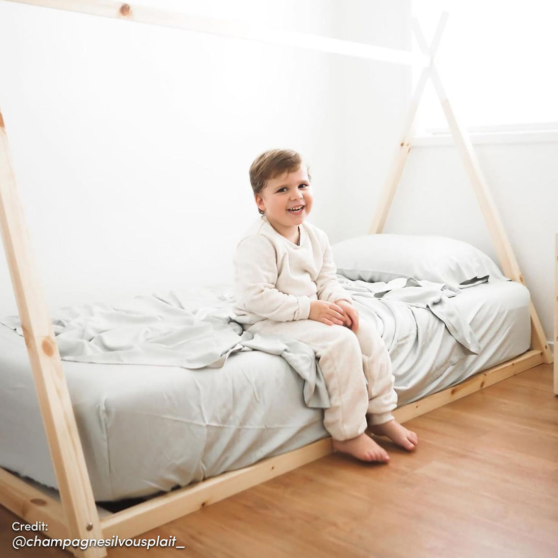 Young boy sits on Helena Kids Single Solid Pine Teepee Bed Frame in Natural, showcasing its low, house-shaped design in a minimalist bedroom with warm wood flooring and white walls.