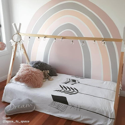 Helena Kids Single Solid Pine Teepee Bed Frame in Natural, featuring a wooden A-frame with globe string-lights, low mattress, fur-style pillows, stuffed elephant, cloud cushion, and dreamcatcher, on a wood-tone floor.