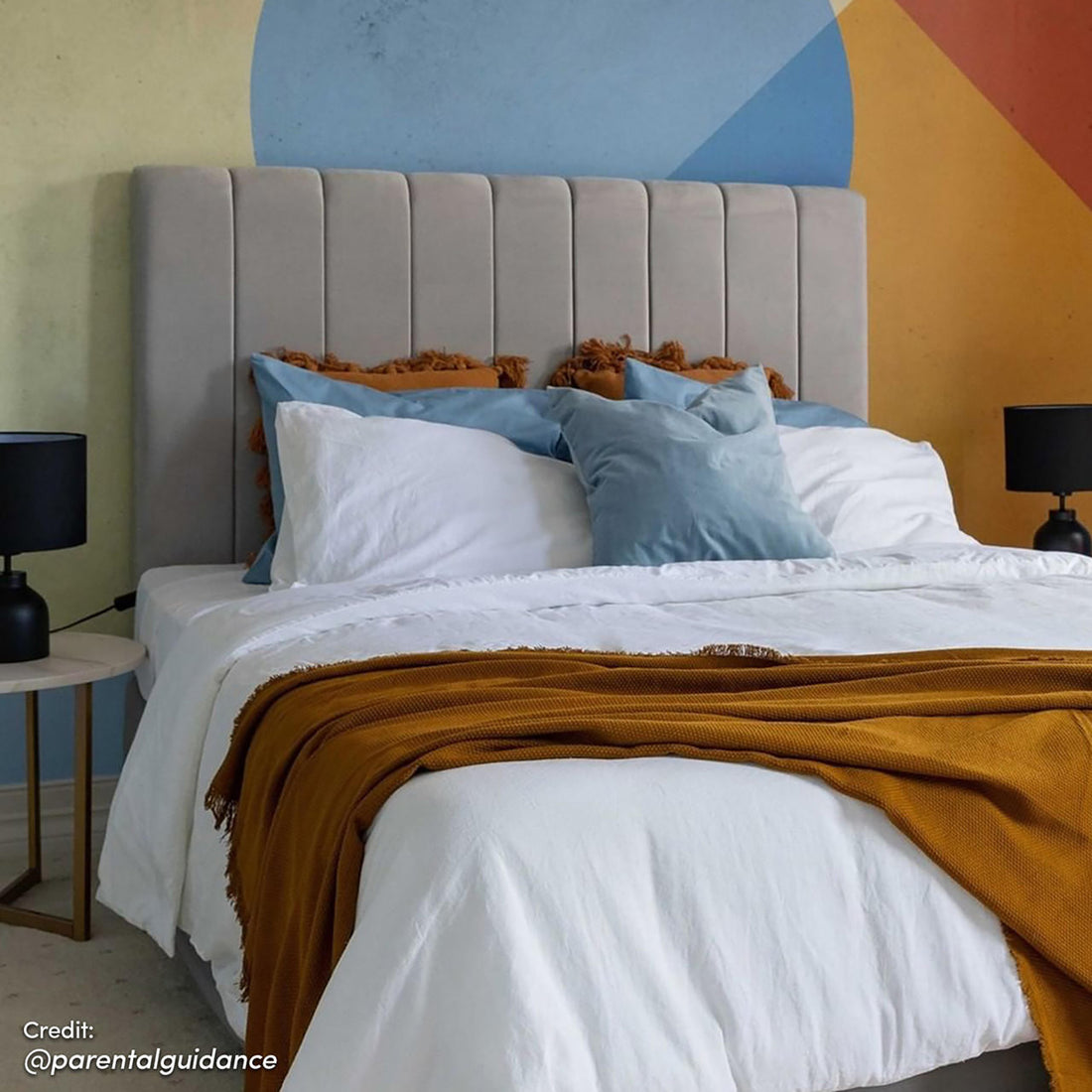 Gratiana Queen Velvet Fabric Headboard in Slate, features light gray channeled upholstery, enhancing a modern bedroom with white bedding, rust-orange throw, accent pillows, bedside tables, and geometric backdrop wall.