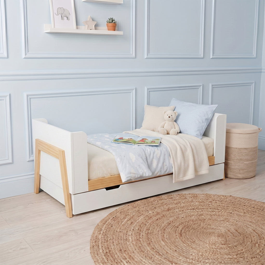White and natural wood baby cot with pull-out storage drawer, shown with bedding and a teddy bear in a nursery setting