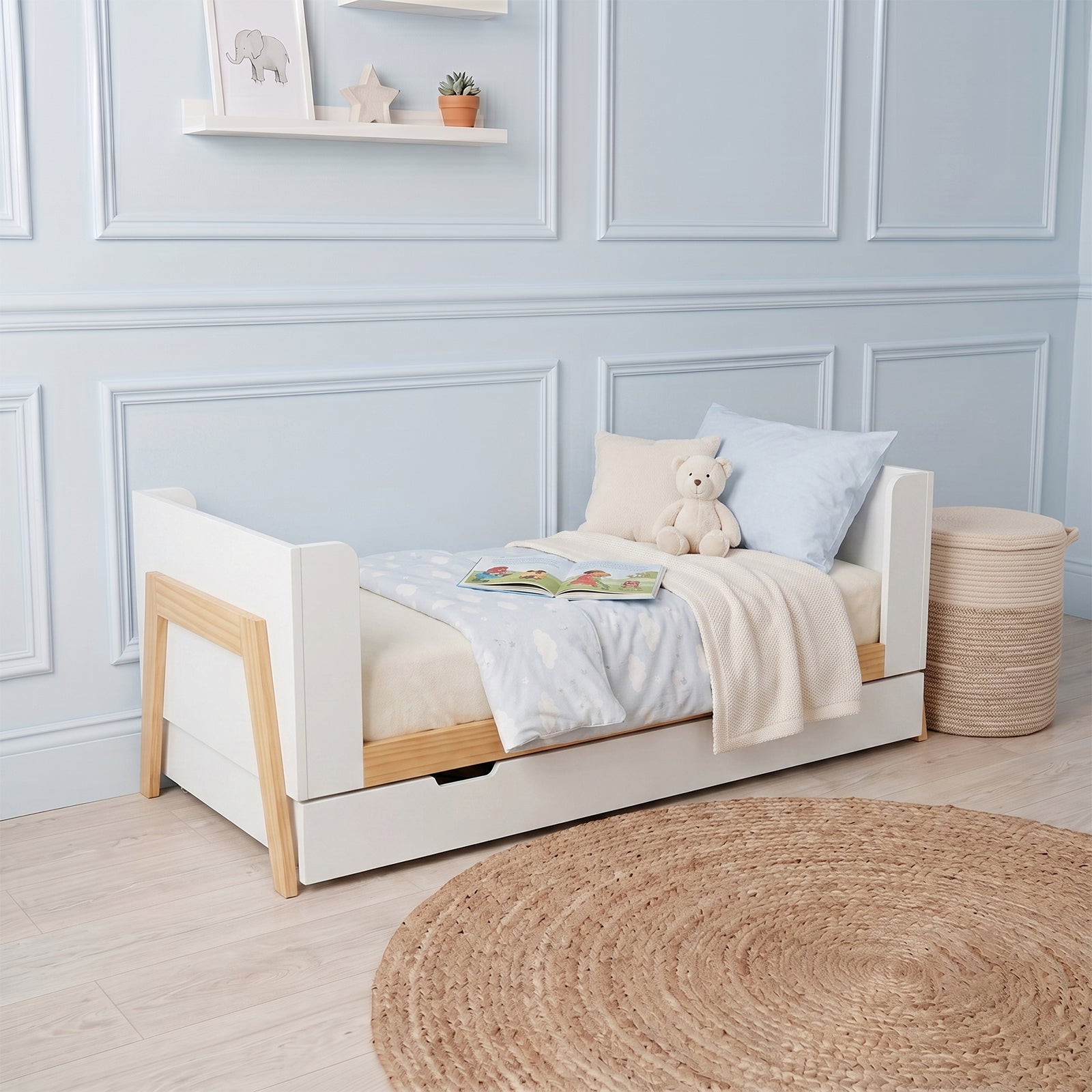White and natural wood baby cot with pull-out storage drawer, shown with bedding and a teddy bear in a nursery setting