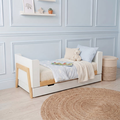 White and natural wood baby cot with pull-out storage drawer, shown with bedding and a teddy bear in a nursery setting