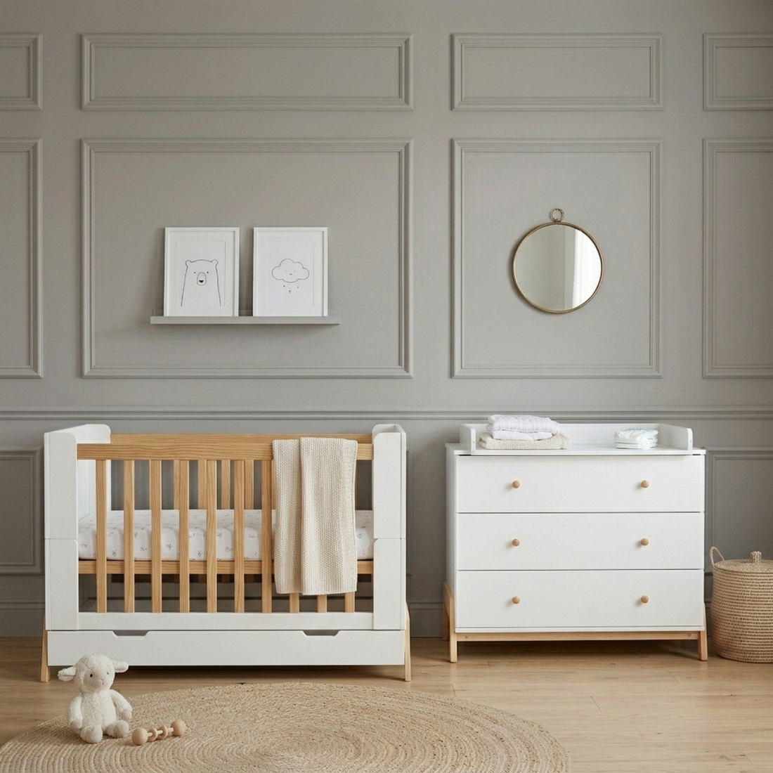 White and natural wood baby cot with storage drawer beside white chest of drawers in grey paneled nursery