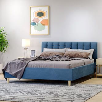 Kelly Velvet Bed Frame - Queen (Ocean Blue) featuring a plush, rectangular headboard with vertical tufting, complemented by elegant tapered legs against a bedroom setting with a lamp and picture frame.