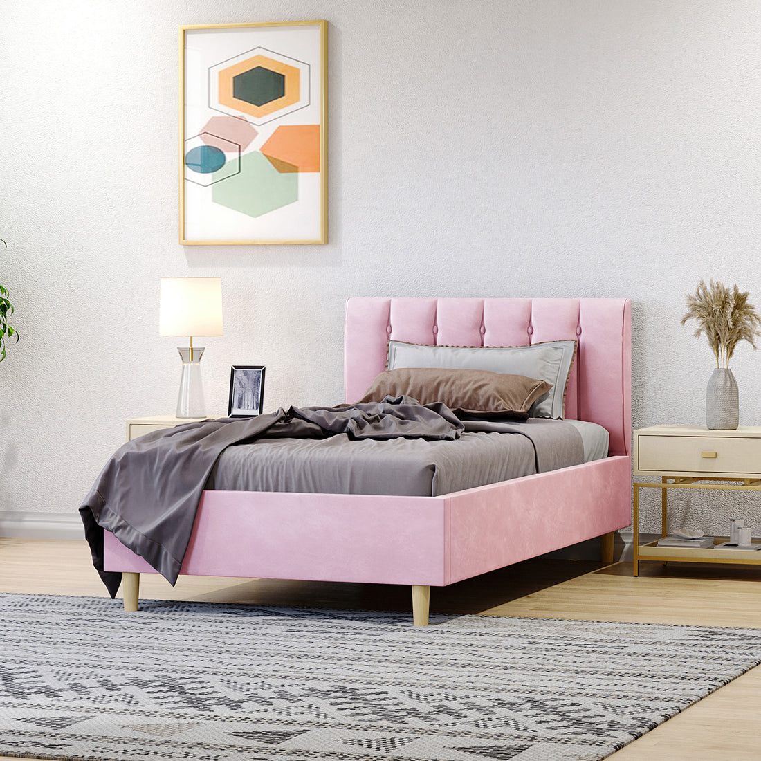 Kelly Single Velvet Fabric Bed Frame in Pink, featuring a vertically tufted headboard, is styled with gray bedding. Flanked by light-wood side tables and decor, all set atop a textured area rug.