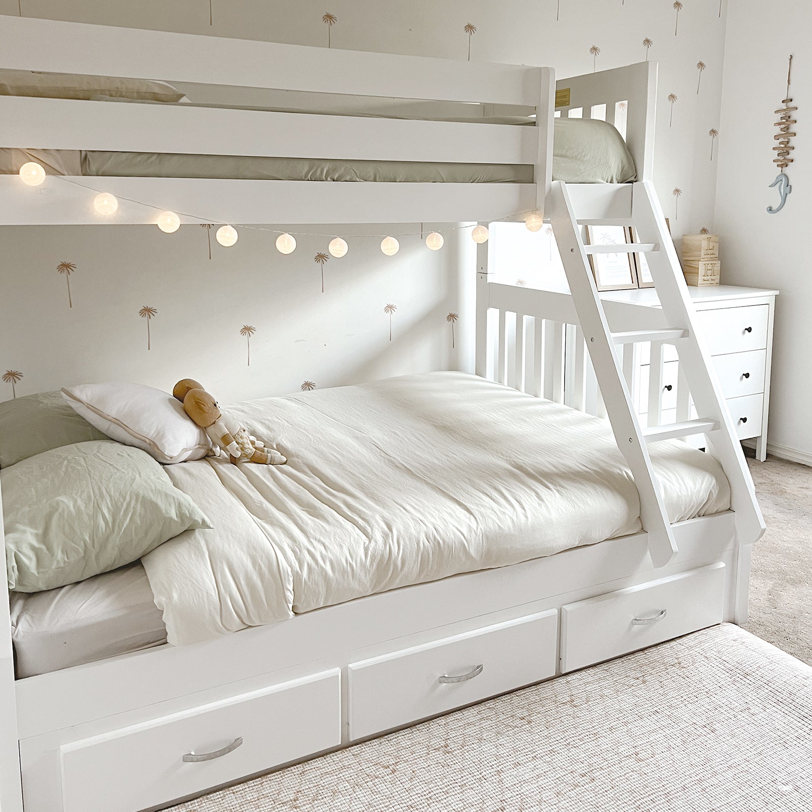 Kourtney Hardwood Triple Bunk Bed with Drawers in White, featuring a twin-over-full layout, pull-out storage drawers, and a slanted ladder, styled with soft bedding and globe lights in a cozy bedroom corner.